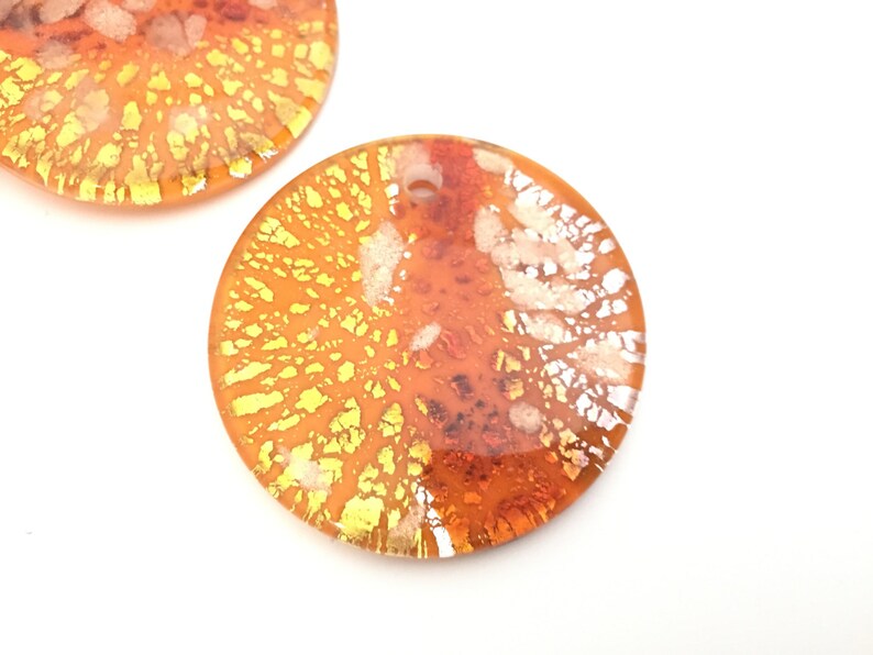 May include: Two orange and yellow dichroic glass pendants with a circular shape. The pendants have a hole at the top for stringing. The glass has a textured, layered appearance with gold and white accents. The pendants are set against a white background.