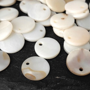 May include: A pile of white and cream colored round shell beads with a hole in the center. The beads are arranged in a random pattern on a black surface.