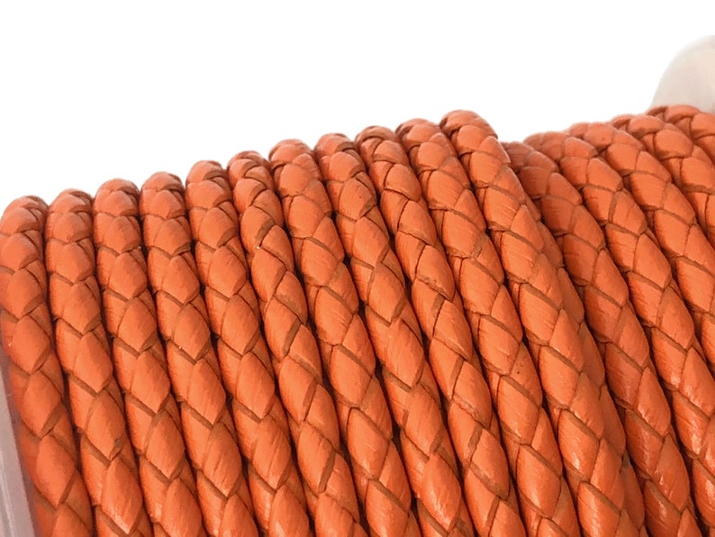 Puede incluir: Primer plano de un carrete de cord&oacute;n de cuero trenzado naranja. El cord&oacute;n est&aacute; enrollado apretado y tiene un acabado liso y pulido.