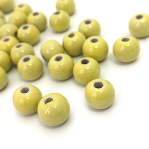 May include: A collection of pale yellow ceramic beads with a glossy finish. Each bead is round with a hole running through the center. The beads are scattered on a white surface, suitable for jewelry making or crafting.
