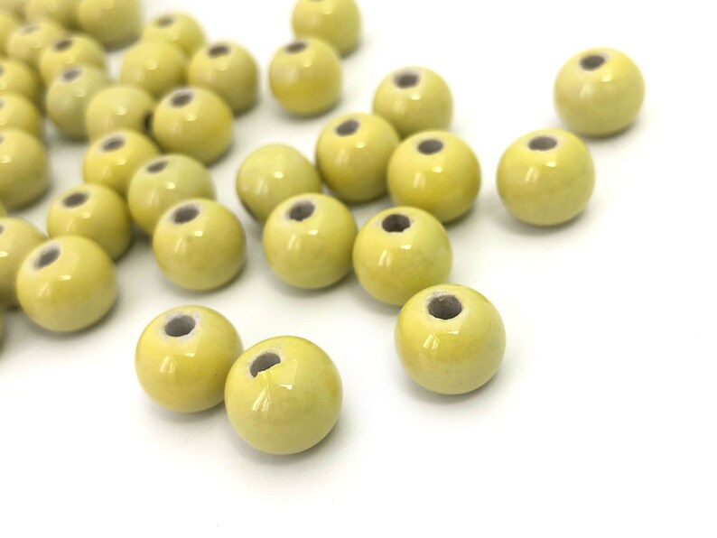 May include: A collection of pale yellow ceramic beads with a glossy finish. Each bead is round with a hole running through the center. The beads are scattered on a white surface, suitable for jewelry making or crafting.