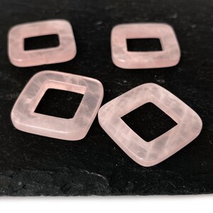 Peut inclure: Quatre perles carr&eacute;es de couleur rose avec un trou au centre. Les perles sont sur une surface noire.