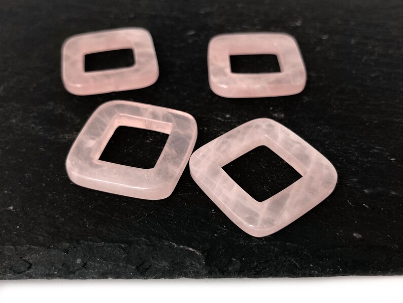 Peut inclure: Quatre perles carr&eacute;es de couleur rose avec un trou au centre. Les perles sont sur une surface noire.