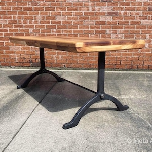 May include: A rectangular wooden table with a light brown surface. The table has black metal legs. The legs are curved and have a unique design. The table is set against a red brick wall.