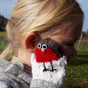 May include: A child wearing a gray sweater with a knitted robin finger puppet on their hand. The robin is red, white, and brown with black eyes and a black beak.