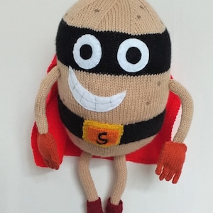 Puede incluir: Un juguete de patata de punto que lleva una capa roja, una máscara negra y un cinturón con una 'S' amarilla. La patata tiene una cara sonriente y está sentada con las piernas cruzadas.