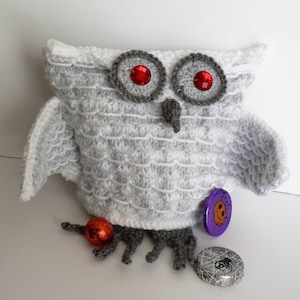 May include: White crocheted owl with red eyes and grey accents. The owl is holding a small orange pumpkin and has a purple and silver candy wrapper on its leg.