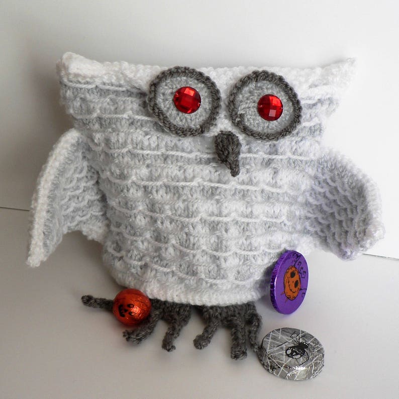 May include: White crocheted owl with red eyes and grey accents. The owl is holding a small orange pumpkin and has a purple and silver candy wrapper on its leg.