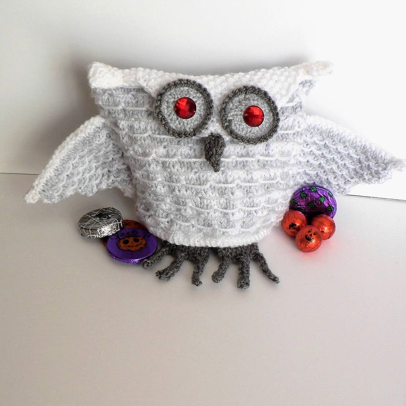 May include: White crocheted owl with red eyes and grey feet. The owl is holding a variety of Halloween sweets in its wings.