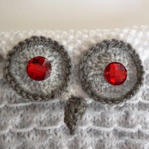 May include: A white and grey knitted owl with red gem eyes. The owl is crocheted and has a textured surface.