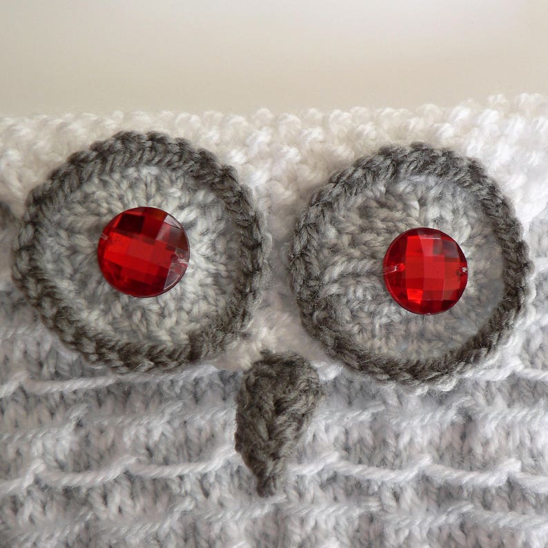 May include: A white and grey knitted owl with red gem eyes. The owl is crocheted and has a textured surface.