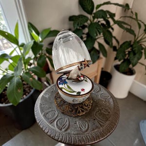 May include: A decorative egg-shaped trinket box with a clear glass top and a white porcelain base. The base features painted blue iris flowers and a small red ladybug. Inside, a small white cat figurine sits on a platform with a gold star.
