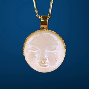K&ouml;nnte beinhalten: Eine goldene Kettenkette mit einem runden Anh&auml;nger, der ein wei&szlig;es, geschnitztes Gesicht zeigt. Der Anh&auml;nger hat einen goldenen Rand.