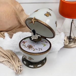 Może przedstawiać: Biała i złota ceramiczna pozytywka z figurką szczeniaka w środku. Pozytywka ma pokrywę na zawiasach ze złotymi śladami łap i złotą gwiazdą. Podstawa ma kwiatowy wzór i ślady łap.