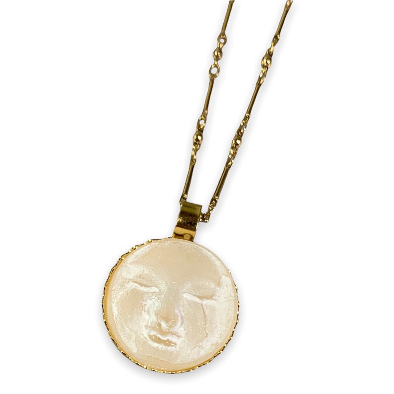 K&ouml;nnte beinhalten: Eine goldene Kettenkette mit einem runden Anh&auml;nger, der ein wei&szlig;es Gesicht mit geschlossenen Augen zeigt. Der Anh&auml;nger hat einen goldenen Rand.