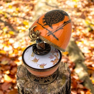 May include: An orange decorative egg-shaped container with a black owl design and a small owl figurine inside. The container has a gold-colored metal hinge and base, and is set on a tree stump. The interior of the container has a floral design.
