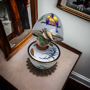 Può includere: Un carillon decorativo a forma di uovo con coperchio incernierato, con un disegno di uccelli dipinti nei toni del blu, giallo e rosa. La base ha un motivo floreale e una piccola statuina di uccello. Il carillon è su una superficie beige.