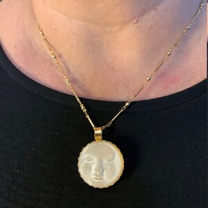 K&ouml;nnte beinhalten: Eine goldene Kettenkette mit einem runden Anh&auml;nger, der ein wei&szlig;es Kamee mit dem Gesicht einer Frau zeigt. Das Kamee ist in einen goldenen Rahmen gefasst.