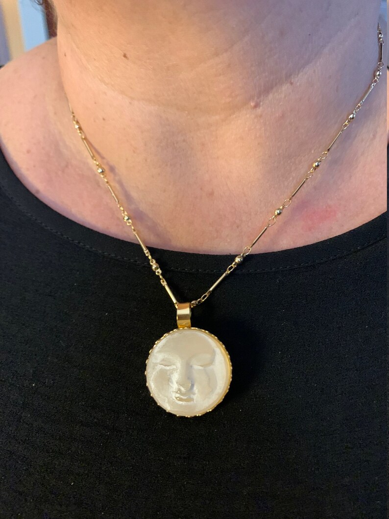 K&ouml;nnte beinhalten: Eine goldene Kettenkette mit einem runden Anh&auml;nger, der ein wei&szlig;es Kamee mit dem Gesicht einer Frau zeigt. Das Kamee ist in einen goldenen Rahmen gefasst.