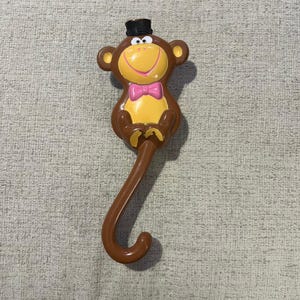 Op de afbeelding: Een bruine, aapvormige parapluhandgreep met een geel gezicht, een zwarte hoge hoed en een roze vlinderdas. De handgreep is gebogen en heeft aan het uiteinde een haak. De aap lacht.