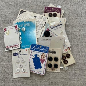 May include: A selection of vintage buttons on cards. The buttons are in various colours and shapes, including white, black, and pearl. Some cards have text such as "Cool White" and "Luckyday".