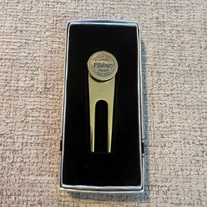 Puede incluir: Una herramienta dorada para reparar divots de golf con el logotipo de Pillsbury Bakery, presentada en una caja negra con un borde a rayas. La herramienta tiene un diseño de horquilla de dos puntas y una parte superior circular.