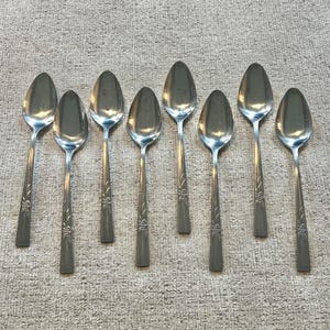 Puede incluir: Un juego de ocho cucharas de metal plateado dispuestas en dos filas. Cada cuchara presenta un cuenco ovalado liso y pulido, y un mango recto decorado con un sutil motivo floral grabado cerca de la base del tallo. El metal tiene un acabado brillante.
