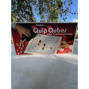 May include: A vintage Scrabble Quip Qubers Cross Sentence Board Game box. The box is red with the game title in white and red. The game board is visible with wooden letter tiles and a person writing on a notepad. The background is a natural outdoor setting.