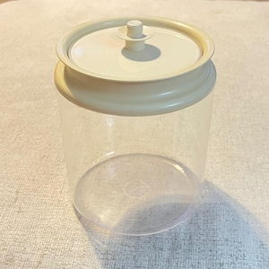 Può includere: Un contenitore cilindrico trasparente con un coperchio in plastica color crema. Il coperchio presenta un bordo circolare rialzato e un piccolo pomello centrale che funge da maniglia. Il corpo del contenitore è completamente trasparente.