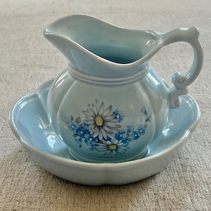 May include: A light blue ceramic pitcher and matching basin set. The pitcher features a curved handle and a floral decal of white daisies and small blue flowers on the front. The basin has a scalloped edge and a smooth, glossy finish throughout.