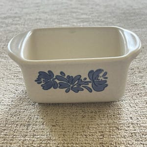 Puede incluir: Una fuente rectangular de cerámica blanquecina con asas redondeadas. La fuente presenta un estampado floral azul en la parte delantera. El interior es de un tono más claro de blanquecino. La fuente se utiliza probablemente para hornear o servir.