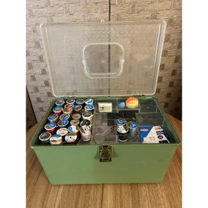 Può includere: Una scatola da cucito verde vintage con un coperchio in plastica trasparente, aperta per rivelare rocchetti di filo, aghi e altri accessori per cucire. La scatola è rettangolare con una chiusura e cerniere in metallo.