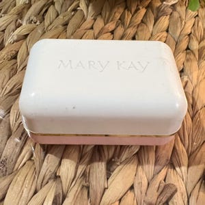 May include: A rectangular cosmetic case in white and pink, with the embossed text "MARY KAY" on the lid. The base is pink with a gold trim. The case is set against a woven, natural-toned background.