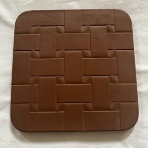 May include: A square, brown, flat object with rounded corners featuring a raised, repeating basket-weave pattern across its entire surface. The design consists of interlocking rectangular segments that create a textured, grid-like appearance.