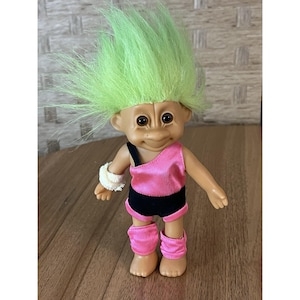 May include: A vintage Troll doll with green hair, wearing a pink leotard, black shorts, and pink leg warmers. The doll has a tan complexion, large brown eyes, and a smiling expression. A white wristband is on one arm.