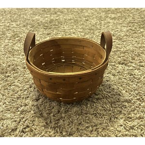 Può includere: Un cesto rotondo in legno intrecciato con finitura marrone medio, bordo rinforzato e due manici verticali fissati ai lati. Il cesto è costruito con listelli di legno piatti sovrapposti in un design classico, robusto e aperto nella parte superiore.