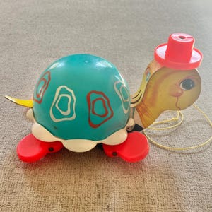 May include: A vintage pull-toy turtle featuring a teal shell with red and white abstract patterns, a yellow head with a red hat, and red feet. The toy includes a yellow pull string attached to the front, and the Fisher-Price logo is visible.