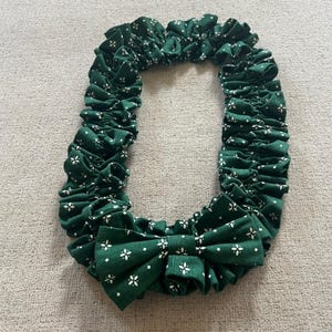 May include: A dark green fabric accessory featuring a repeating white floral pattern. The item is constructed with a gathered, ruffled design forming an elongated oval shape, finished with a prominent, structured bow at the bottom centre.