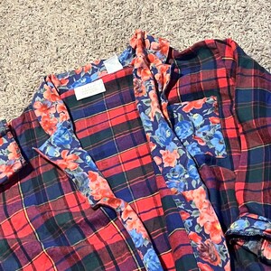 May include: A long-sleeved button-down shirt featuring a red, navy blue, and dark green plaid pattern. The garment includes a contrasting floral print trim in shades of blue, orange, and red along the collar, front placket, and chest pocket.