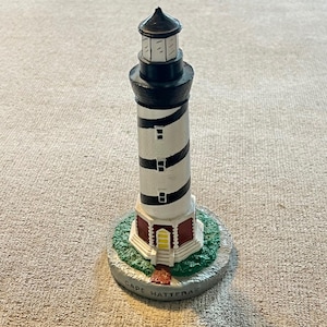 May include: A miniature lighthouse figurine featuring a white tower with black spiral stripes, a black lantern room, and a red base with a yellow door. The base is textured with green grass and the text CAPE HATTERAS is printed on the front edge.