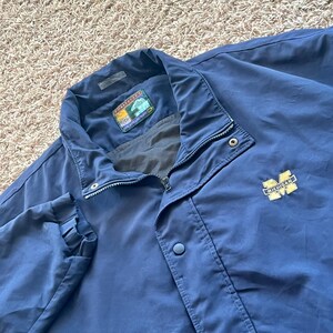 May include: A navy blue windbreaker jacket featuring a high collar, a front zip closure with a snap-button placket, and a yellow embroidered Michigan block M logo on the chest. The interior neck label displays the Forrester's brand logo.
