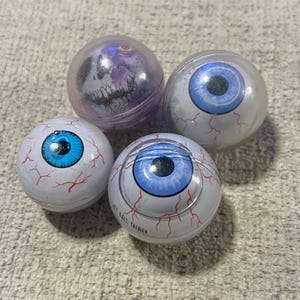 Op de afbeelding: Vier bolvormige speeltjes die lijken op oogballen. Drie zijn wit met blauwe irissen en rode aders. Eén is een doorschijnende paarse bol met een schedelafbeelding. De tekst JET BALL TAIWAN staat gedrukt op de zijkant van een wit oogbal-speeltje.