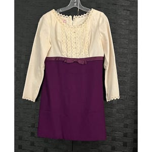 Könnte beinhalten: Cremefarbenes Kleid mit langen Ärmeln und lila Rock. Das Oberteil hat Spitzendetails und eine kleine Schleife an der Taille. Das Kleid hängt an einem weißen Kleiderbügel.
