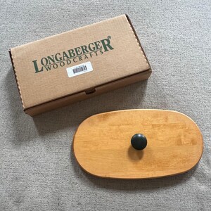 May include: A rectangular cardboard box with the green text LONGABERGER WOODCRAFTS printed on the lid, accompanied by an oval-shaped light brown wooden lid featuring a centred, smooth, black circular knob handle for a container or basket.
