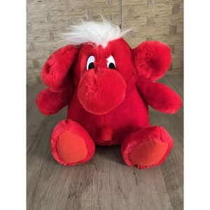 May include: A plush red stuffed animal with large ears, a round nose, and white hair. The toy has orange accents on its feet and is sitting on a wooden surface. The toy is a cartoon-like character.
