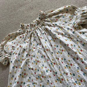 May include: A white blouse featuring a small-scale floral pattern in red, yellow, and blue, accented by tiny blue dots. The garment has a gathered, ruffled neckline and short, puffed sleeves with elasticized, ruffled cuffs, creating a loose silhouette.