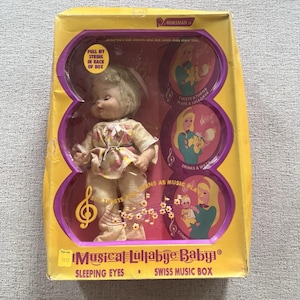 Pode incluir: Boneca vintage "Musical Lullabye Baby" na embalagem original. A boneca está vestida com uma roupa estampada floral e tem olhos de dormir. A caixa é amarela e roxa com ilustrações e texto.