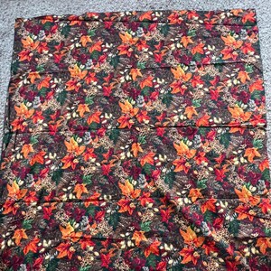 May include: A rectangular fabric piece featuring a dense autumn-themed pattern. The design displays vibrant orange, red, and yellow maple leaves interspersed with green pine needles, brown pinecones, and small acorns against a dark brown background.