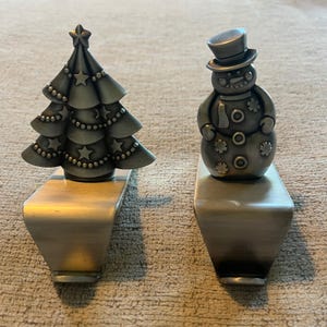 May include: Two silver-toned metal stocking holders. One features a tiered Christmas tree decorated with stars and beaded garlands. The other is a snowman wearing a top hat, adorned with snowflake and circular accents. Both have flat bases.