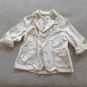 Può includere: Un blazer beige chiaro con colletto a revers classico e due tasche frontali. La giacca ha maniche lunghe con polsini ripiegati e chiusura con un solo bottone. Il tessuto sembra essere un materiale morbido e strutturato.